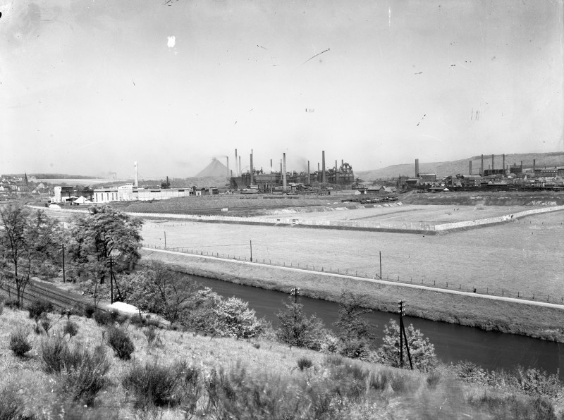 Auf dem scharz-weiß Foto ist eine Landschaft zu sehen: Ein begrühnter kleiner Hügel im Vordergrund, ein schmaler Fluss, dahinter die Industriegebäude der Völklinger Hütte, Berge am Horizont.